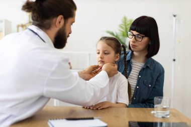Deneyimli erkek doktor annesiyle otururken beyaz kız lenf bezlerini inceliyor. Anne yakından izliyor, ilgi ve ilgi gösteriyor..