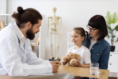 Kafkasyalı erkek doktor, klinikte genç kız ve annesiyle sağlığı tartışıyor. Kız oyuncak ayıyı tutuyor, danışma sırasında rahatlık yaratıyor..