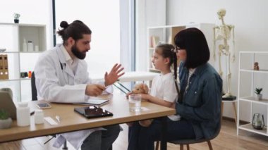 Kafkas erkek doktor klinikte alnına dokunarak genç kızın ateşini ölçüyor. Anne, beyaz kadın, kızlarının omzunun yanında oturuyor..