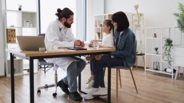 Erkek doktor, muayenehanedeki genç kız ve anneyle etkileşimde. Görüntüde Kafkas erkek doktor, örgülü beyaz kız ve Kafkas kadın anne yer alıyor..
