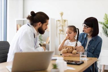 Kafkas erkek doktor, annesi için reçete yazarken klinikte öksüren beyaz kızı muayene ediyor. Kız rahatlamak için oyuncak ayıyı tutuyor. Etkileşim bakımı, sağlık danışmanlığını somutlaştırır.