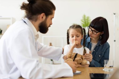 Kafkas erkek doktor, endişeli Kafkas anne için reçete yazarken klinikte öksüren genç beyaz kızı muayene ediyor. Tıbbi danışmanlık sağlık hizmetlerinde bakım ve kararlılığı vurguluyor.