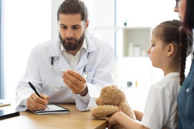 Kafkas erkek doktor oyuncak ayı tutan küçük beyaz kızın ateşini kontrol ediyor. Sahne, pediatri kliniğinde ilgili ve ilgili sağlık hizmetlerini resmediyor.