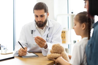 Kafkasyalı erkek doktor, klinikte oyuncak ayıyı tutan genç kıza danışmanlık yapıyor. Doktor, hastaya notlar yazarken, şefkatli sağlık hizmetlerinin altını çizerken odaklanır..