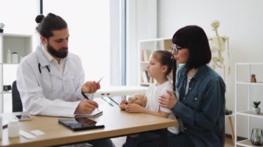 Beyaz önlüklü beyaz erkek doktor, klinikte annesinin kucağında oturan küçük hasta, beyaz bir kızın sıcaklığını ölçüyor. Doktor tıbbi muayene sırasında dijital termometre kullanır..