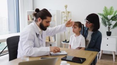 Annesi muayenehanede ona bakarken beyaz erkek doktor hasta genç kızın ateşini ölçüyor. Doktor, kızların alnını inceliyor. Endişe ve bakım gösteriyor..