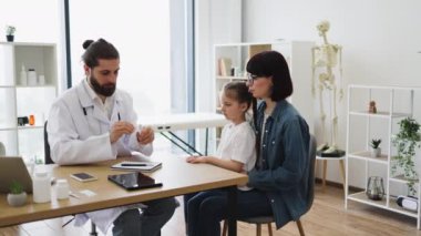 Beyaz erkek doktor, beyaz kızın boğazını tahta spatula kullanarak inceliyor. Annesi, beyaz bir kadın, rahatlık sağlamak için yanında oturuyor..