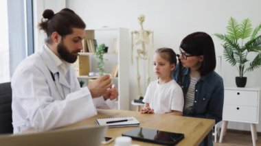 Kafkasyalı erkek doktor, tıbbi spatula kullanarak annesiyle oturan beyaz bir kızın boğazını inceliyor. Modern tıp bürosundaki sahne, sağlık hizmetlerinde sıcaklık ve profesyonel bakım gösteriyor..