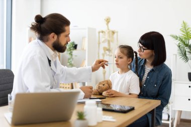 Kafkas erkek doktor annesinin yanında oturan küçük hasta beyaz kızın sıcaklığını ölçer. Doktor doğru okumak için termometreyi kullanırken, anne de oyuncak ayıyı tutan kızı rahatlatıyor.