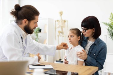 Kafkas erkek doktor klinikteki küçük hasta kızın ateşini ölçüyor. Endişeli görünen çocuk, beyaz kadın ebeveyn tarafından teselli edilir..