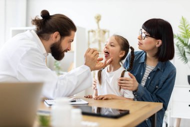 Kafkasyalı erkek doktor, tıbbi spatula kullanarak annesiyle oturan beyaz bir kızın boğazını inceliyor. Modern tıp bürosundaki sahne, sağlık hizmetlerinde sıcaklık ve profesyonel bakım gösteriyor..
