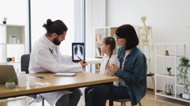 Beyaz erkek doktor akciğer röntgenini genç beyaz kız ve annesiyle klinikte tartışıyor. Doktor beyaz önlük giyiyor, tıbbi görüntüyü göstermek için tablet kullanıyor..
