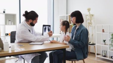Kafkasyalı erkek doktor, parlak klinik ortamında genç bir kız ve anneye akciğer röntgeni gösteriyor. Doktor laboratuvar önlüğü giyer ve gösteri için tablet kullanır. Kız özenli görünüyor ve anne destekleyici bir şekilde oturuyor..