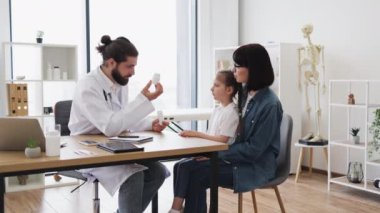 Kafkas erkek doktor hasta kız ve anneye ilaç ve ilaç veriyor. Genç kız modern klinikte annesinin kucağında oturuyor. Profesyonel sağlık hizmetleri ve güveni tasvir ediyor.