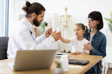 Kafkas erkek doktor, annesiyle klinikte oturan küçük kıza ilaç veriyor. Doktor şişeyi ve su toplamayı uzatırken kız özenli görünüyor. Annem destek olur..