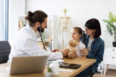 Kafkas erkek doktor, tıbbi klinikte beyaz bir kızı rahatlatmak için oyuncak ayı kullanarak steteskop inceleme tekniği gösteriyor. Annem genç bir kızın yanında oturuyor ve ona destek oluyor.