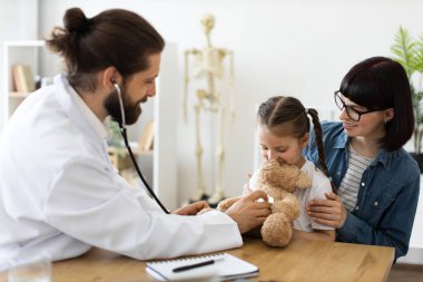 Beyaz erkek doktor, klinikte beyaz bir kızla oyuncak ayıyı kullanarak steteskop muayenesi yapıyor. Gülümseyen destekleyici anne, kızının yanında oturuyor, ziyaret sırasında onu teselli ediyor..