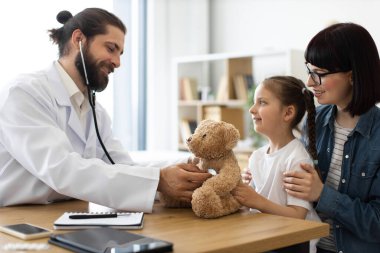 Kafkas erkek doktor, klinikteki genç kız ve annesine tıbbi uygulamaları göstermek için oyuncak ayı kullanarak steteskopla muayene ediyor..