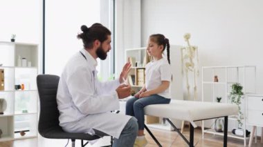 Kafkasyalı erkek doktor, genç kızı tıbbi ortamlarda dil bastırıcı kullanarak muayene ediyor. Sahne, çocuk sağlığını sakin bir mesleki ortamda güvence altına alan doktorla temel pediatrik bakımı yakalıyor