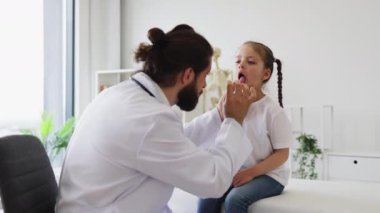 Kafkasyalı erkek doktor, modern tıp kliniğinde dil bastırıcı kullanan bir kızın boğazını inceliyor. Sahne, doktor-hasta etkileşiminin ve teşhisin vurgulandığı profesyonel sağlık ortamını gösteriyor.