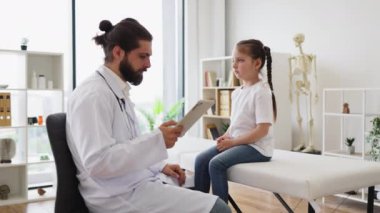 Beyaz erkek doktor, yüksek ateşi olan bir kızı tıbbi kanepede muayene ediyor. Doktor çocuk ateşini ölçmek için termometre kullanıyor. Çocuk sağlığı sağlık hizmetleri ve doktor-hasta ilişkisinin temalarını vurgular