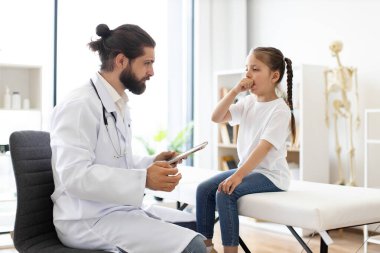 Beyaz erkek doktor öksüren genç kızı muayene ediyor. Sahne, profesyonel bakım ve endişeye odaklı hastane veya klinikte sağlık kontrollerini gösteriyor.