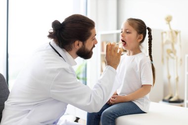 Kafkasyalı erkek doktor, modern tıp kliniğinde dil bastırıcı kullanan bir kızın boğazını inceliyor. Sahne, doktor-hasta etkileşiminin ve teşhisin vurgulandığı profesyonel sağlık ortamını gösteriyor.