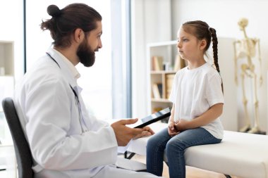 Tıbbi uzman genç beyaz kızı muayene ediyor tıbbi kanepede oturuyor, güneş ışığı odaya akıyor. Doktor, özenli sağlık uygulamalarını gösteren verileri tablete kaydeder..