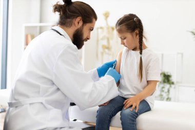 Erkek doktor, beyaz bir kızı şırıngayla tıbbi kanepede aşıladı. Doktor beyaz önlük, eldiven giyer ve kızların kollarına odaklanır. Sahne, sağlık, tıbbi bakım ve aşı sürecini betimliyor.