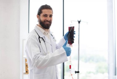 Sakallı bir erkek doktor, laboratuvar önlüğü ve eldiven giyiyor, kan naklini hazırlıyor. Beyaz tenli, 30 'lu yaşlarda, modern tıbbi ekipmanlarıyla parlak hastane ortamında..