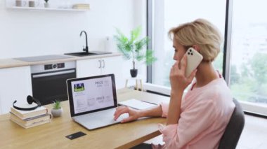 Beyaz bir kadın telefonla konuşurken ve kredi kartı tutarken internetten kozmetik ürünleri sipariş ediyor. Sarı saçlı, yetişkin bir kadın online alışveriş yapıyor ve ev ortamında ödeme yapıyor..