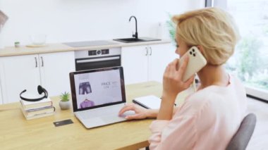 Kafkasyalı bir kadın cep telefonuyla konuşurken dizüstü bilgisayarında giysi dükkanına göz atıyor. Orta yaşlı kadın, modern ve aydınlık mutfaktaki dijital platformu kullanarak kıyafet sipariş ediyor..