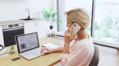 Parlak ev ortamında masasında oturan günlük giysili beyaz bir kadın. İnternetten kıyafet sipariş etmek ve laptop ekranını kontrol etmek için akıllı telefon kullanıyor. Kapalı alan bitkileri ve kitaplar ev atmosferine eklenir.