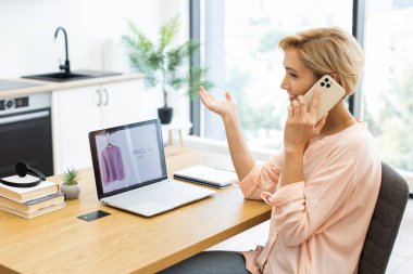 Parlak ev ortamında masasında oturan günlük giysili beyaz bir kadın. İnternetten kıyafet sipariş etmek ve laptop ekranını kontrol etmek için akıllı telefon kullanıyor. Kapalı alan bitkileri ve kitaplar ev atmosferine eklenir.