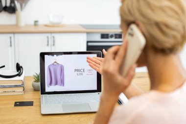 Beyaz bir kadın telefonda konuşurken dizüstü bilgisayar kullanarak internetten kıyafet sipariş ediyor. Senaryo modern dijital alışveriş deneyimini, rahatlığı ve çevrimiçi perakende teknolojisini yansıtıyor.