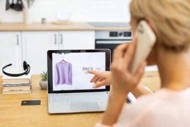 30 'lu yaşlarda beyaz bir kadın telefonda konuşurken dizüstü bilgisayar kullanarak internetten kıyafet sipariş ediyor. Görüntü, teknoloji ve kolaylık odaklı modern yaşam tarzını yakalar.