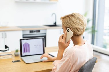 Kafkasyalı bir kadın bilgisayarda gezinirken internetten kıyafet sipariş etmek için akıllı telefon kullanıyor. Parlak, modern bir mutfakta oturuyor, çevrimiçi alışverişin kolaylığını ve çoklu görevini gösteriyor..