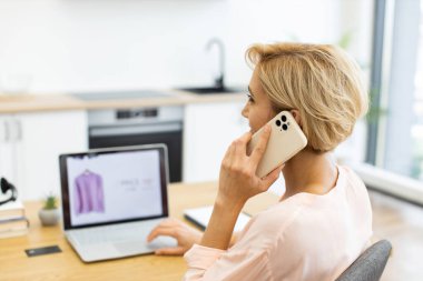 Akıllı telefon ve dizüstü bilgisayar kullanan yirmili yaşlarda beyaz bir kadın. Modern aydınlık ortamda internetten kıyafet sipariş etmek. Çevrimiçi alışveriş ve dijital yaşam tarzı kavramı.