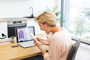 Beyaz bir kadın online alışveriş sorunu hakkında telefonla konuşurken hayal kırıklığını dile getiriyor. Masasında dizüstü bilgisayarında giysi parçalarını gösteriyor. İçişleri Bakanlığı çağdaş alışveriş deneyimini ima ediyor.