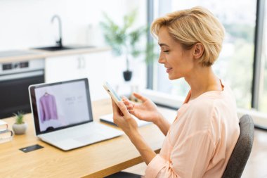 Beyaz kadın telefonda konuşuyor. İnternetten kıyafet alımıyla ilgili endişelerini dile getiriyor. Ev ofisinde dizüstü bilgisayarında web sitesi olan orta yaşlı bir kadın..