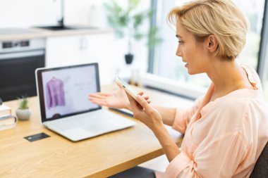 30 'lu yaşlardaki beyaz bir kadın, online giyim mağazasından hayal kırıklığına uğramış bir şekilde evde telefonla konuşuyor. Teslimat veya ürün kalitesinden memnun olmadığını belirten ürün imajını gösteren dizüstü bilgisayara doğru hareket ediyor.
