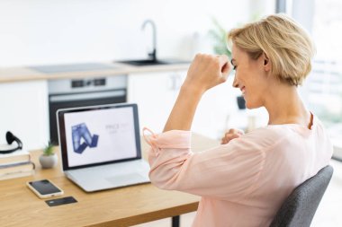 İnternetten alışveriş başarısını kutlayan mutlu beyaz kadın dizüstü bilgisayar kullanıyor. Telefon ve kredi kartıyla konforlu bir evde masasında oturmuş, neşe ve memnuniyeti ifade ediyor..