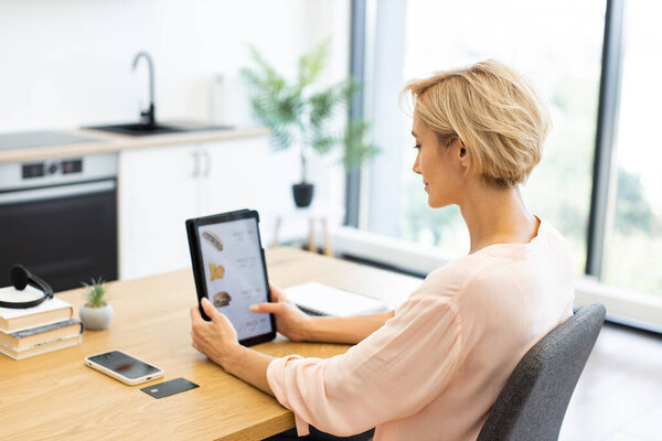 Кавказская женщина использует планшет, чтобы заказать еду онлайн в современных домашних условиях. Случайная сцена показывает цифровые технологии, улучшающие повседневную жизнь.