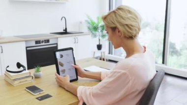 Modern mutfakta tablet kullanarak internetten yemek sipariş ederken mutlu beyaz kadın sevinçle elini kaldırıyor. Hevesli ve memnun görünüyor, menü seçeneklerini gösteren bir cihazla yemek masasında oturuyor..