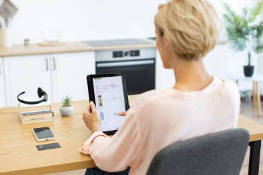 Modern mutfakta otururken kozmetik ürünleri sipariş etmek için tablet kullanan beyaz bir kadın. Tablet ekranda dağıtım sitesinde kozmetik ürünler gösteriliyor. Samimi ayarlar cep telefonu ve kredi kartı içerir.