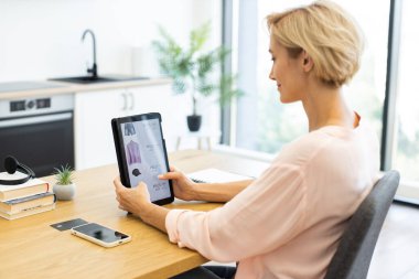 Caucasian woman browsing online store using tablet, seated at home office desk. She appears focused on selecting clothing from delivery website. clipart