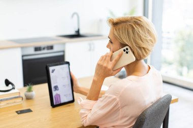 Tabletteki giyim sitesine bakarken akıllı telefon kullanan beyaz bir kadın. Modern mutfakta masa başında oturmak, teslimat hizmeti ile internetten kıyafet sipariş etmek..