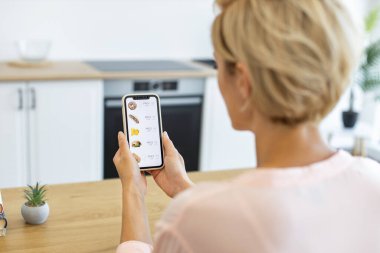 Beyaz bir kadın internet sitesinden yemek sipariş etmek için akıllı telefon kullanıyor. Modern mutfakta oturuyor, mobil cihazda çeşitli yiyecek seçeneklerini araştırıyor..