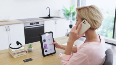 30 'lu yaşlarda beyaz bir kadın evde tablet kullanarak internetten kıyafet sipariş ediyor. Parlak, modern bir mutfak ortamında ahşap bir masada oturuyor. Teknoloji ve e-ticaret üzerinde duruluyor.