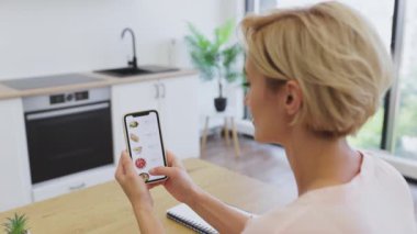 Beyaz bir kadın internet sitesinden yemek sipariş etmek için akıllı telefon kullanıyor. Modern mutfakta oturuyor, mobil cihazda çeşitli yiyecek seçeneklerini araştırıyor..